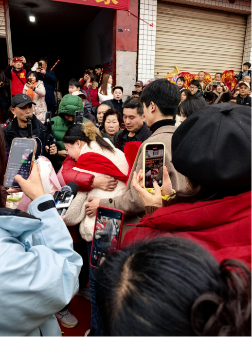 温馨纪实画面：白发苍苍的父母紧握女儿双手，三人身着新中式礼服站在庭院前微笑，背景悬挂‘团圆’书法横幅